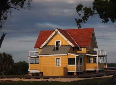 [This two-story house is a much smaller version of the Instone house in that it also has porches on the first two levels and is the same color scheme. This building has much steeper-pitched roofing which is light red in color. From this back-side view, it appears the building is on level ground. A lot of white smeary clouds fill the blue sky around the building.]